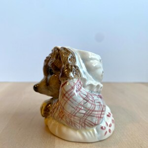 May include: A ceramic figurine of a hedgehog wearing a red and white checked shirt and a white bonnet. The hedgehog is sitting on a white cushion with red polka dots.