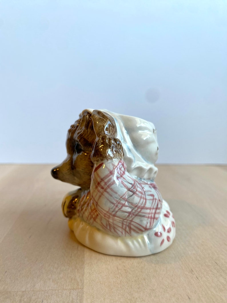 May include: A ceramic figurine of a hedgehog wearing a red and white checked shirt and a white bonnet. The hedgehog is sitting on a white cushion with red polka dots.