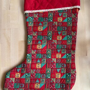 May include: A quilted Christmas stocking with a red cuff and a patterned body. The body features a patchwork design with red, green, and gold squares, triangles, and rectangles. The cuff is a solid red with a white floral trim.