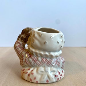 May include: A ceramic figurine of a girl wearing a red and white checked dress and a white bonnet. The figurine has a small opening on the top of the head, suggesting it may be a planter or a vase.
