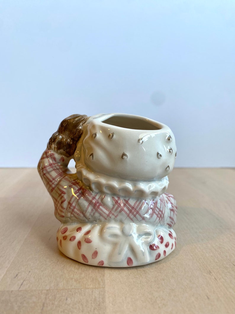 May include: A ceramic figurine of a girl wearing a red and white checked dress and a white bonnet. The figurine has a small opening on the top of the head, suggesting it may be a planter or a vase.