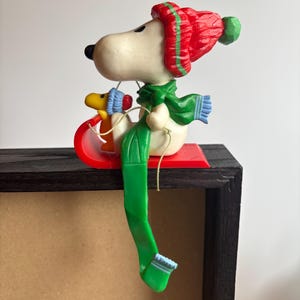 May include: A Snoopy figurine wearing a red hat and green scarf, sitting on a red sled with Woodstock. The figurine is perched on a dark wooden ledge. The hat has green stripes and a green pom-pom.