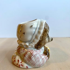 May include: A ceramic figurine of a small animal wearing a pink and white dress and a white bonnet. The figurine has a brown body and a white face. The figurine is sitting on a wooden surface.