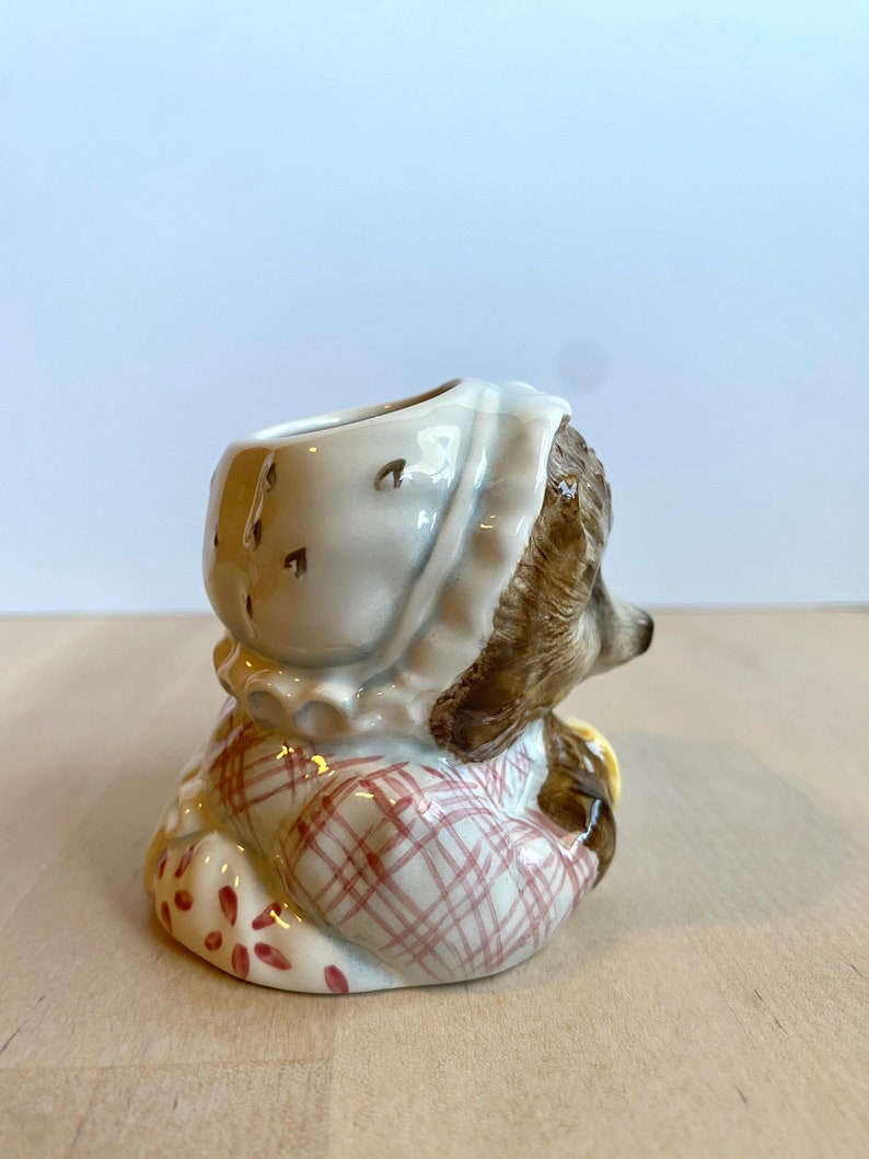 May include: A ceramic figurine of a small animal wearing a pink and white dress and a white bonnet. The figurine has a brown body and a white face. The figurine is sitting on a wooden surface.