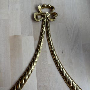 May include: A gold-colored metal decorative piece, featuring a bow-shaped top with two rope-like strands extending downwards. The piece has a shiny finish and is likely used for decorative purposes.