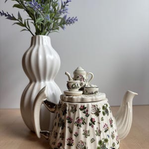 May include: A cream-colored teapot with a floral pattern, featuring a miniature teapot and teacups on top. A white vase with lavender sprigs is in the background. The teapot has a curved handle and a bird-shaped spout.
