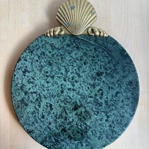 May include: A round, green marble serving tray with a decorative brass seashell handle. The tray has a speckled pattern and a polished surface. The seashell handle is detailed with small blue accents.