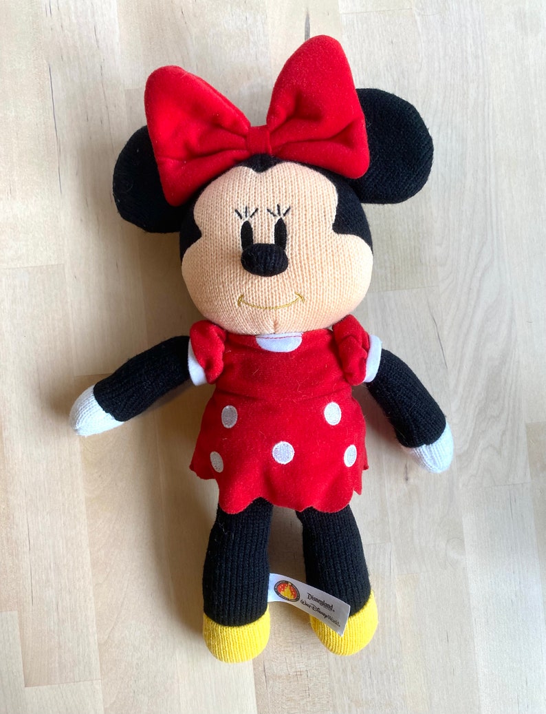 May include: Knitted Minnie Mouse plush toy wearing a red dress with white polka dots and a red bow. The toy has a yellow tag that says "Disneyland."