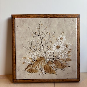 May include: Square ceramic tile with a floral design in a wooden frame. The artwork features brown and white flowers, leaves, and berries on a beige background. The wooden frame is a medium brown color.