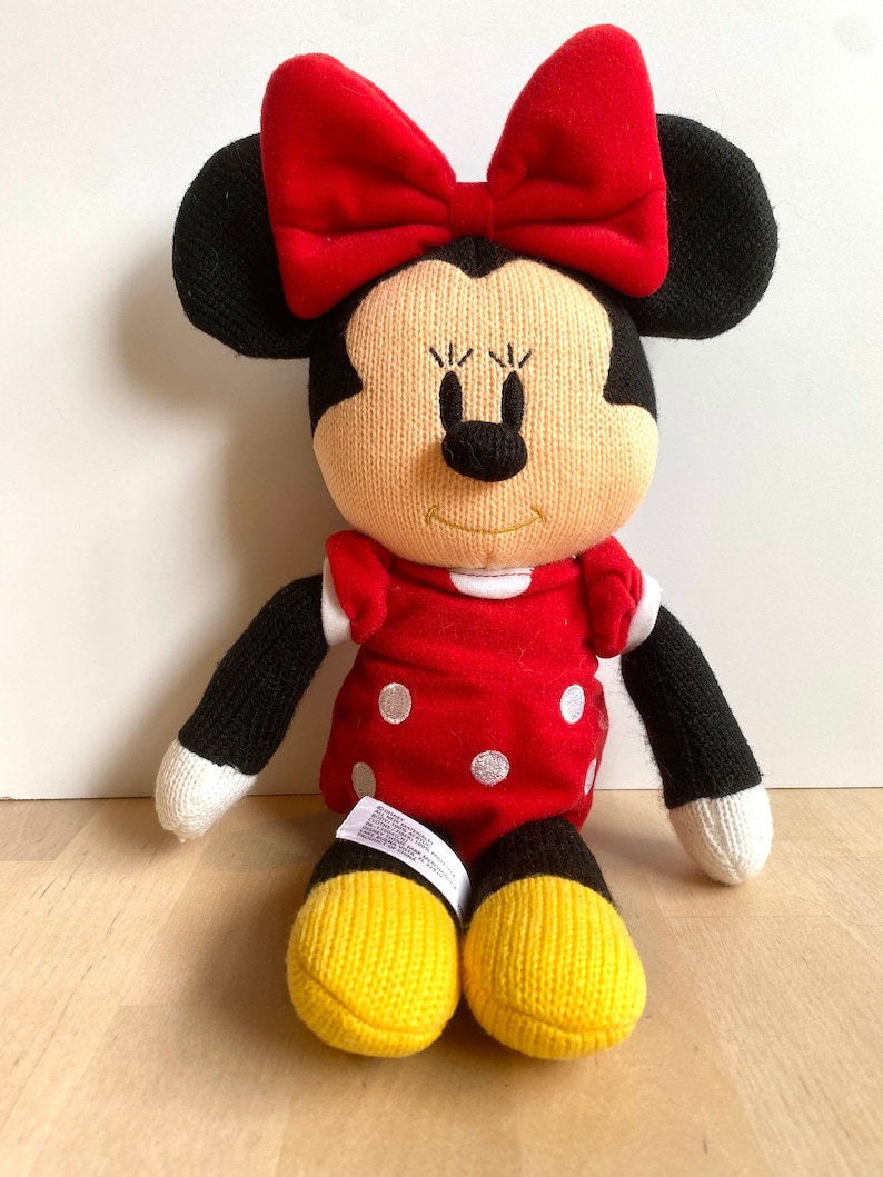 May include: A red and black knitted plush toy of Minnie Mouse. She is wearing a red dress with white polka dots and a red bow.
