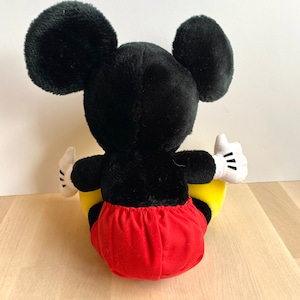 以下が含まれることがあります： 黒と赤のぬいぐるみミッキーマウスが、黄色のぬいぐるみ三日月の上に座っています。
