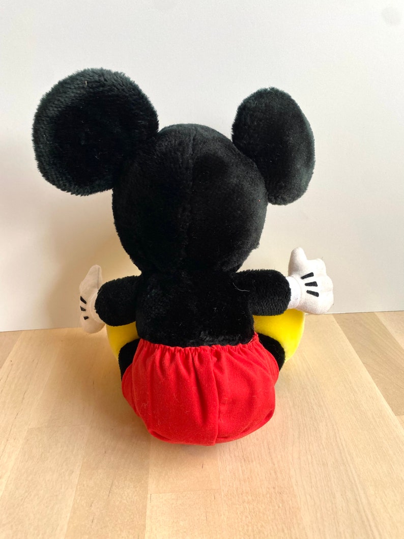 以下が含まれることがあります： 黒と赤のぬいぐるみミッキーマウスが、黄色のぬいぐるみ三日月の上に座っています。