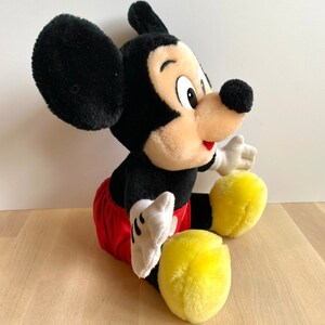 以下が含まれることがあります： 足を組んで座っているミッキーマウスのぬいぐるみ。赤いショートパンツを履いており、黒い毛皮に黄色の靴を履いています。
