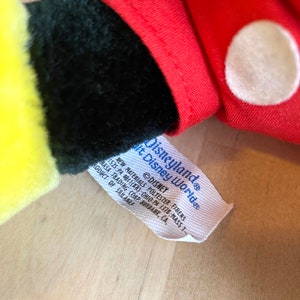 以下が含まれることがあります： 「Disneyland&reg; It Disney World&reg; Disney」と「New Materials Polyester Fibers 1525:PA 40111KR): Ohio PA 1378 Mass T NASA Trading Corp Burbank, CA, Product of Sri Lanka」と書かれた黒い文字の白いタグ。