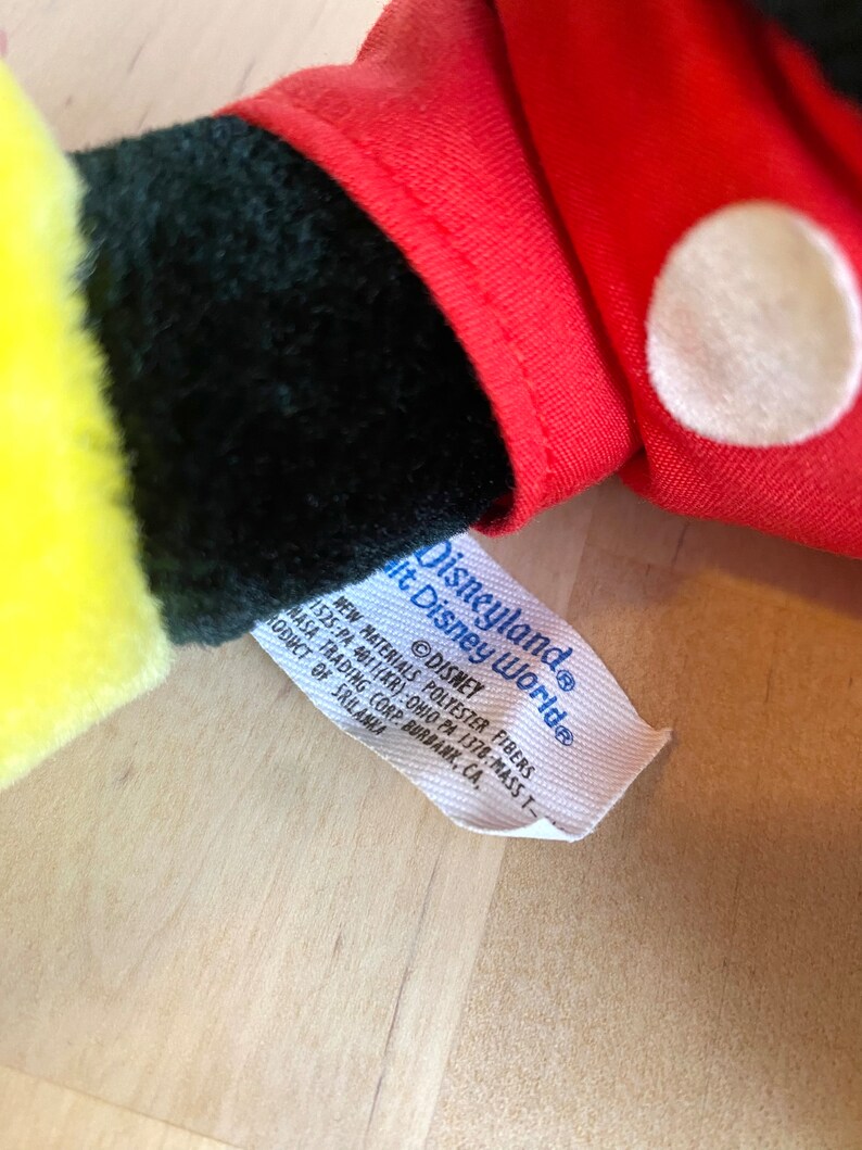 以下が含まれることがあります： 「Disneyland&reg; It Disney World&reg; Disney」と「New Materials Polyester Fibers 1525:PA 40111KR): Ohio PA 1378 Mass T NASA Trading Corp Burbank, CA, Product of Sri Lanka」と書かれた黒い文字の白いタグ。