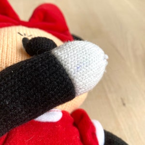 May include: A knitted plush toy of a cartoon character wearing a red dress, black arms, and a white face. The toy has a red bow on its head and a white glove on one hand.