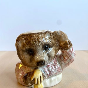 May include: A ceramic figurine of a hedgehog wearing a red and white checked shirt and holding a yellow object. The figurine is brown and white and has a detailed face.