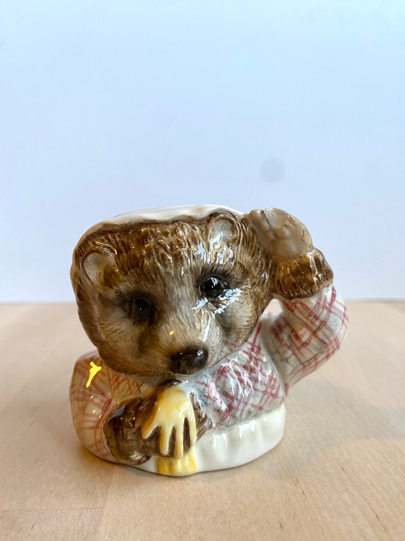 May include: A ceramic figurine of a hedgehog wearing a red and white checked shirt and holding a yellow object. The figurine is brown and white and has a detailed face.