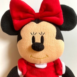 May include: A plush toy of Minnie Mouse wearing a red dress with white polka dots and a red bow. She has black ears and a black nose.