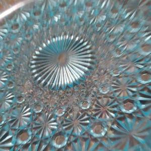 May include: A light blue glass plate featuring a pressed pattern of radiating starbursts and circular indentations. The design radiates from a central sunburst motif, creating a textured surface with geometric shapes across the entire circular dish.