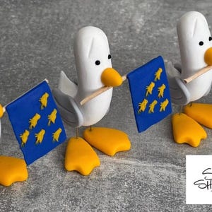 Puede incluir: Tres figuras caprichosas de gaviota con cuerpos blancos, alas grises y picos y patas amarillas. Cada una sostiene una bandera azul con detalles amarillos. Las figuras están sobre una superficie gris. El logotipo "Seagull Shenanigans" está en la esquina inferior derecha.