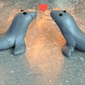 May include: Two grey seal figurines, each with black eyes and whiskers, facing each other with a red heart above them. The image includes the text "Seagull Shenanigans" in a small white box.