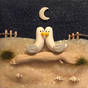 Könnte beinhalten: Ein skurriles Diorama mit zwei weißen Möwenfiguren mit grauen Flügeln und gelben Schnäbeln, die sich küssen. Sie sitzen auf einem Treibholzstamm, mit einem Mond und einem sternenklaren Nachthimmel als Hintergrund. Kleine Muscheln und Grasbüschel vervollständigen die Strandszene.