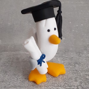 May include: A white bird figurine, wearing a black mortarboard and holding a scroll tied with a blue ribbon. It has orange feet and a yellow beak. The figurine is a graduation theme, on a grey surface.