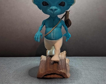 May include: A blue figurine of a creature with a white mushroom-shaped hat, white pants, and a small bag. The figurine is standing on a wooden base. The creature has black eyes and is carrying a small snail on its back.