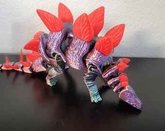 May include: A purple and teal dinosaur toy with red spikes and a long tail. The dinosaur has a textured surface and is made of plastic.