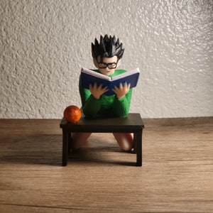 May include: A green and black figurine of a person sitting on a small black table, reading a blue book. A small orange sphere with red stars sits on the table.