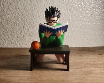 May include: A green and black figurine of a person sitting on a small black table, reading a blue book. A small orange sphere with red stars sits on the table.