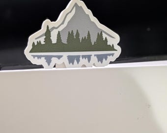 以下が含まれることがあります： 山脈のデザインが施されたステッカー。白い縁取り、灰色の山、緑の森、そして下部に青い反射があります。このデザインは、山岳風景の様式化された表現です。