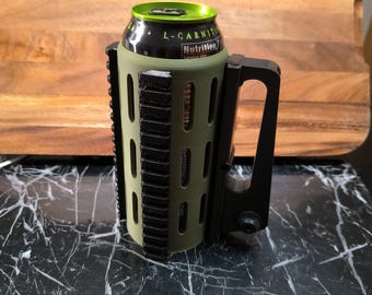 May include: An olive green can cooler with a black handle and a black rail system. The cooler holds a black can with green lettering and a green lid. The background is a wooden cutting board.