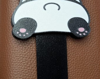 以下が含まれることがあります： パンダの形をした気まぐれなブックマーク。ブックマークは、ピンク色の肉球が付いた白黒のパンダのデザインが特徴です。パンダの後ろ姿が見え、黒いストラップが下に向かって伸びています。背景は質感のある茶色の表面です。
