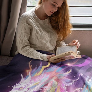 Puede incluir: Una manta morada oscura con una colorida ilustraci&oacute;n de un drag&oacute;n con detalles en blanco, rosa, amarillo y azul. La cabeza del drag&oacute;n es prominente. La manta est&aacute; sobre una persona leyendo un libro.