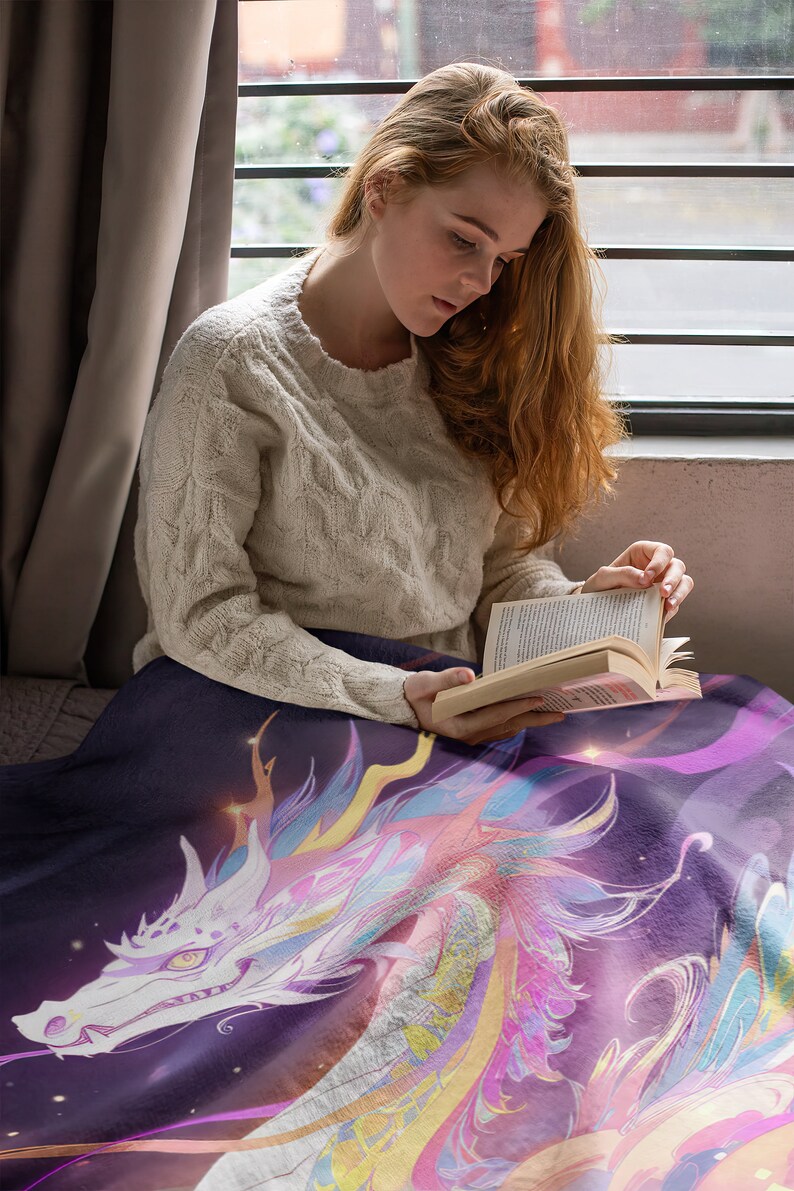 Puede incluir: Una manta morada oscura con una colorida ilustraci&oacute;n de un drag&oacute;n con detalles en blanco, rosa, amarillo y azul. La cabeza del drag&oacute;n es prominente. La manta est&aacute; sobre una persona leyendo un libro.