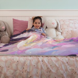 Puede incluir: Una colorida manta con un dise&ntilde;o de unicornio en tonos morados, rosas y amarillos. La manta est&aacute; colocada sobre una cama con ropa de cama con estampado de cisnes. La imagen incluye peluches y almohadas, creando una escena acogedora y atractiva.
