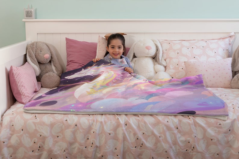 Puede incluir: Una colorida manta con un dise&ntilde;o de unicornio en tonos morados, rosas y amarillos. La manta est&aacute; colocada sobre una cama con ropa de cama con estampado de cisnes. La imagen incluye peluches y almohadas, creando una escena acogedora y atractiva.