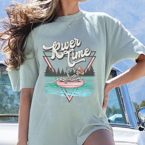 Puede incluir: Una camiseta de gran tamaño de color azul claro con un esqueleto que lleva un sombrero de vaquero y sostiene una bebida en un flotador tubular en un río. El texto "River Time" está en una fuente retro sobre el esqueleto.