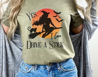Puede incluir: Camiseta verde oliva con un diseño temático de Halloween. El diseño presenta una bruja en una escoba con un gato negro, murciélagos y el texto "Yes, I can drive a stick." La camiseta se combina con pantalones cortos de mezclilla.