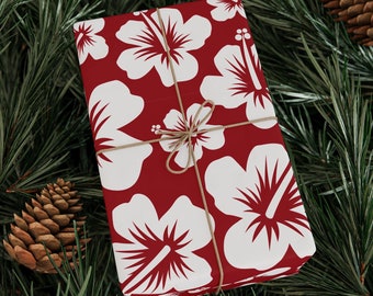 May include: A red gift box with white hibiscus flowers wrapped with twine. The box is sitting on a bed of green pine needles.