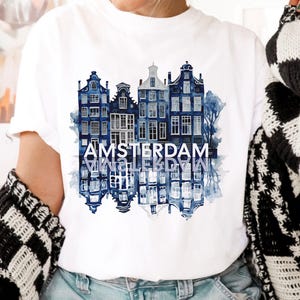 Może przedstawiać: Biała koszulka z akwarelowym wzorem domów na kanałach Amsterdamu w odcieniach niebieskiego. Słowo "AMSTERDAM" jest wydrukowane na biało, z lustrzanym odbiciem poniżej. Koszulka jest noszona z niebieskimi dżinsami i czarno-białym swetrem.