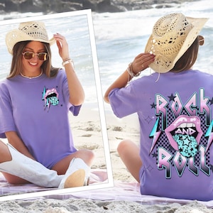 May include: A lilac-colored t-shirt with a graphic design featuring a mouth with a star-filled tongue and lightning bolts. The back of the shirt displays the words "ROCK AND ROLL" with a checkered background and a similar mouth graphic.
