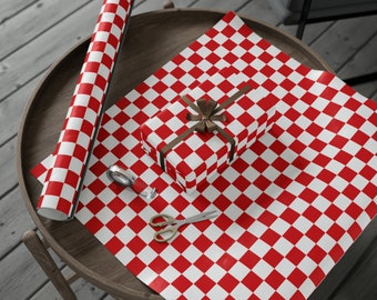 May include: A roll of red and white chequered wrapping paper and a wrapped gift with a brown ribbon sit on a wooden table. A pair of gold scissors and tape are also on the table. The gift is wrapped in the same chequered paper.