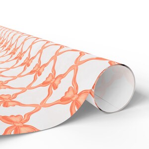 Può includere: Rotolo di carta da regalo con un motivo ripetuto di nastri e fiocchi color corallo su sfondo bianco. I fiocchi sono distanziati in modo uniforme e creano un design a forma di griglia. La carta è parzialmente srotolata.