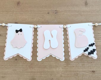 May include: A decorative banner featuring three scalloped-edge pennants strung on a white ribbon with wooden beads. The first pennant shows a light pink ghost with a black bow, the second a white letter N, and the third a white letter E with black bats.