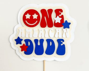 Puede incluir: Un adorno para tarta en capas con el texto ONE AMERICAN DUDE en rojo, blanco y azul. La palabra ONE incluye una cara sonriente roja con ojos de estrella. El diseño está decorado con estrellas y montado sobre un palito de madera.