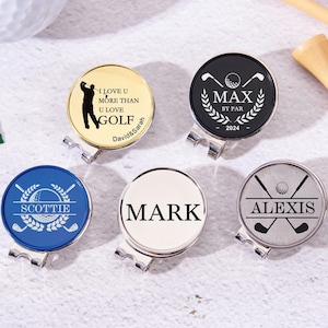 May include: Five different golf ball markers with personalized engravings. The markers are round and feature various designs, including a golfer, crossed golf clubs, and a golf ball with a wreath. The markers are made of metal and have a silver, gold, black, white, and blue finish.