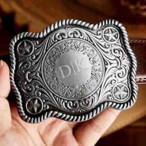 May include: Silver belt buckle with an ornate design featuring a circle with the initials "DK" in the center. The buckle has a rope-like border and three five-pointed stars.